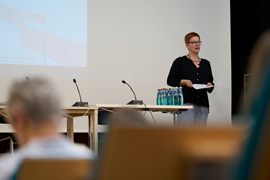 Johanna Pink steht auf der Bühne und spricht. Im Vordergrund sieht man Hinterköpfe des Publikums.